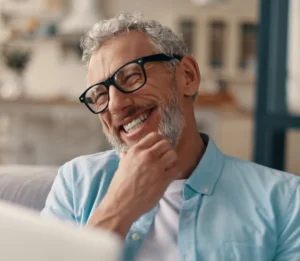 Hombre mayor sonriente con pelo y barba canosos que lleva gafas negras y camisa azul claro, sentado en el interior y mirando una pantalla.
