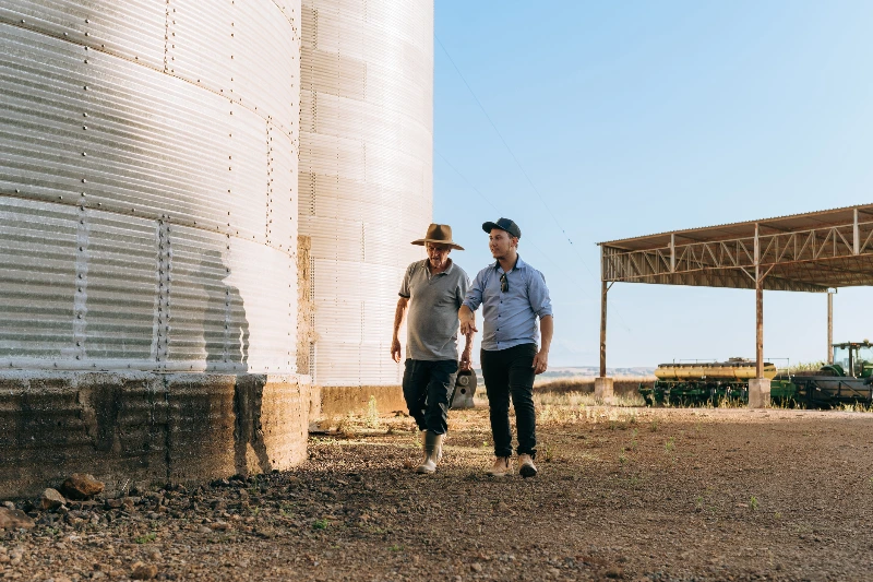 Two men, one older wearing a hat and the younger one in a cap, walking together next to large silos on a farm during the day.