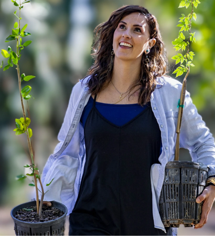 A smiling young woman with long curly hair stands outdoors holding small tree saplings, wearing a blue top and white jacket. Green leaves are visible in the foreground and background.