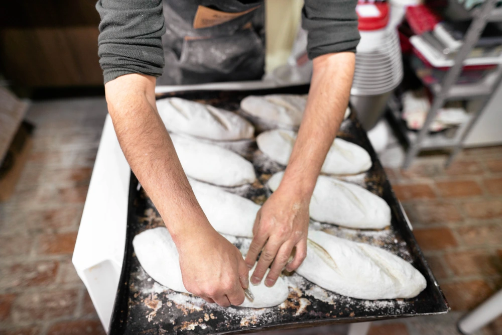 A baker's hands preparing loaves of bread on a floured baking tray in a bakery kitchen.