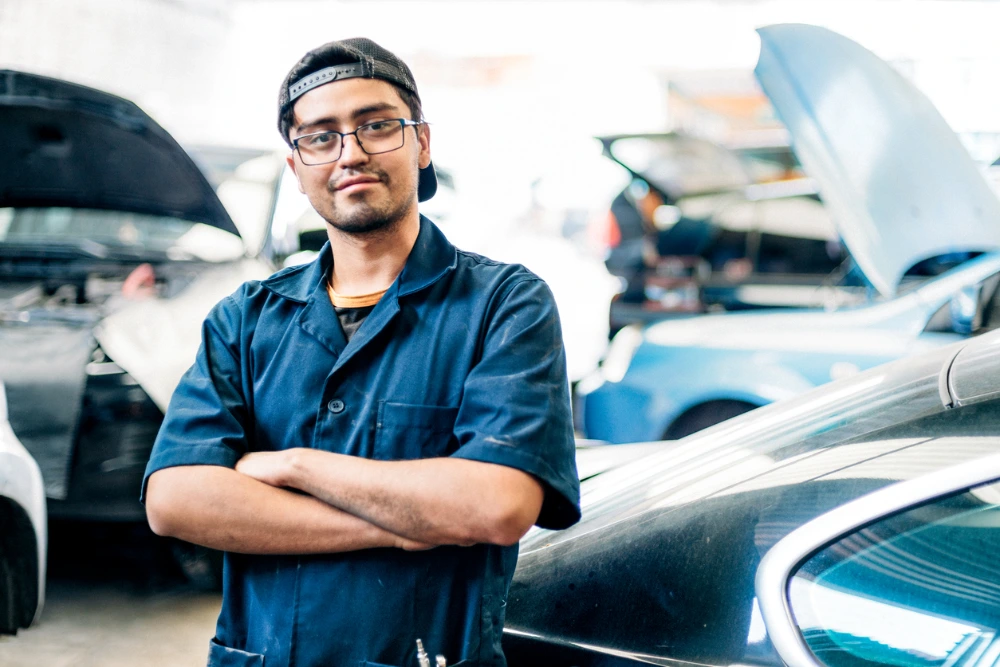 A confident mechanic with crossed arms standing in a car repair shop with vehicles and open hoods in the background.