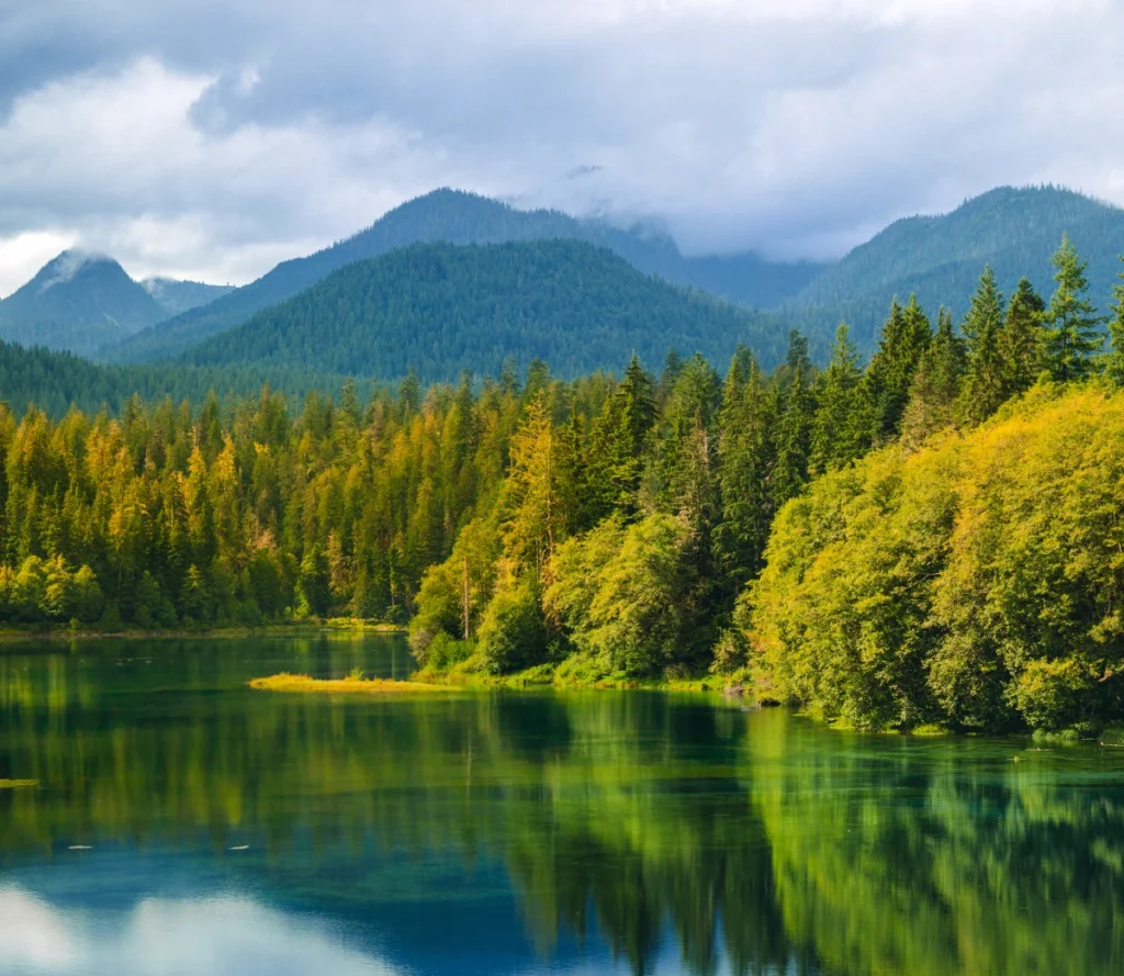 Scenic mountain lake surrounded by dense green forest with reflections in the water under a cloudy sky.