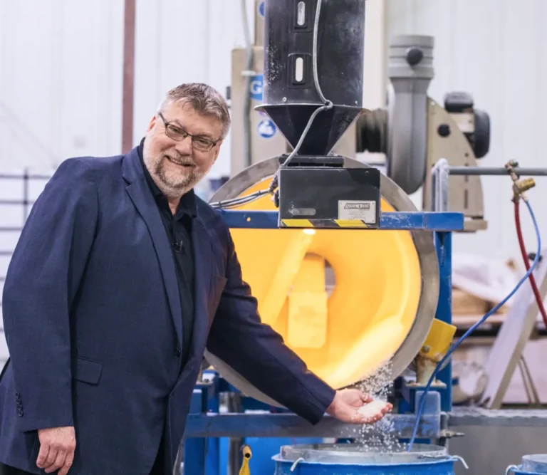 Hombre trajeado sonriendo mientras sostiene bolitas de plástico procedentes de una máquina industrial en una planta de fabricación.