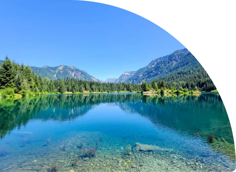 Clear mountain lake reflecting forested slopes and peaks under a bright blue sky