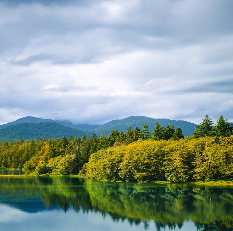 Scenic view of a calm lake reflecting lush green forest and mountains under a cloudy sky.