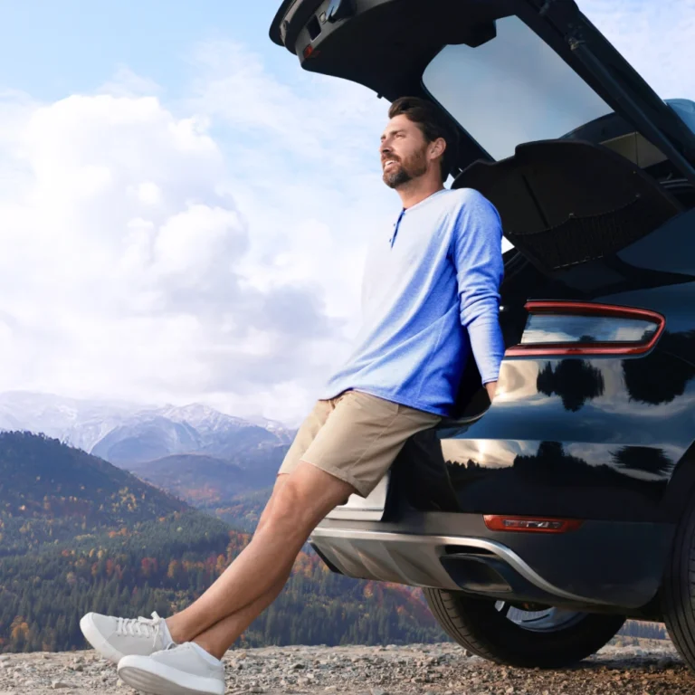 Man relaxing at the back of an SUV parked on a scenic mountain overlook, wearing a light blue long-sleeve shirt, khaki shorts, and white sneakers, with a backdrop of forested hills and snow-capped peaks under a partly cloudy sky.