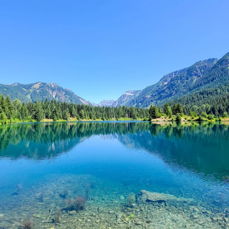 Clear mountain lake reflecting forested slopes and peaks under a bright blue sky